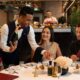 A waiter in a formal vest is presenting a bottle of champagne to a smiling woman seated at a white-clothed dinner table with two other diners in an upscale restaurant.