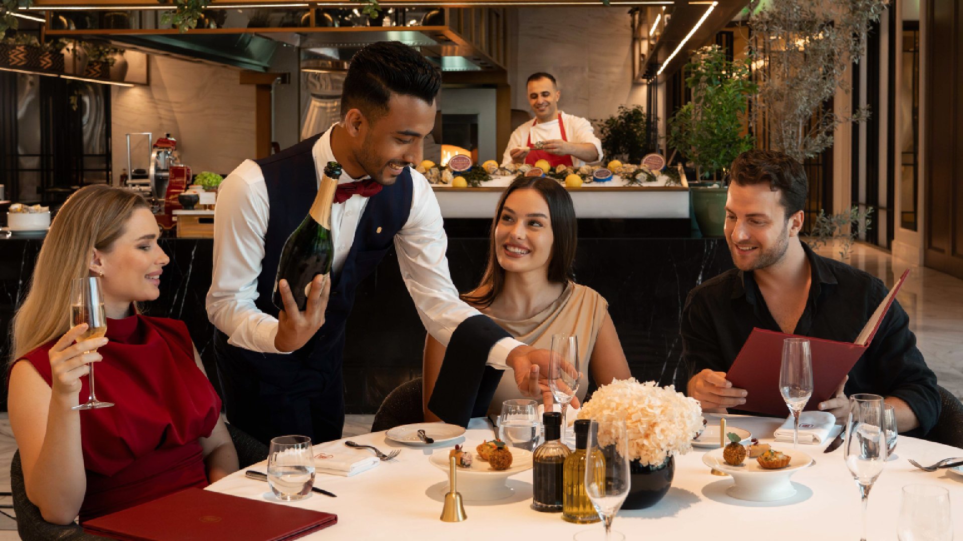 A waiter in a formal vest is presenting a bottle of champagne to a smiling woman seated at a white-clothed dinner table with two other diners in an upscale restaurant.