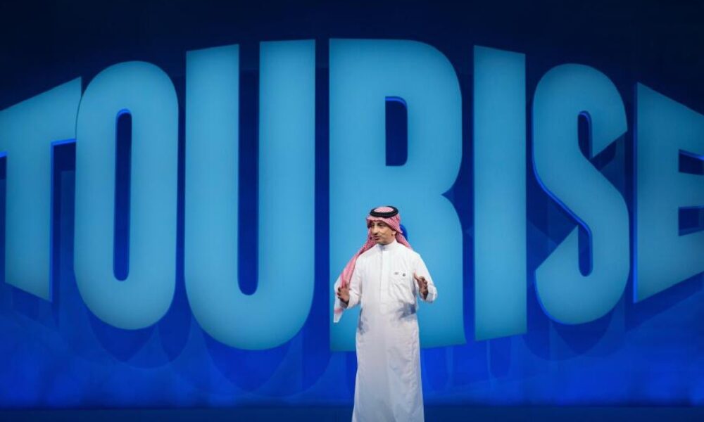 Speaker in traditional Gulf attire presenting on stage with large blue "TOURISE" text backdrop