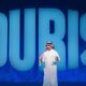 Speaker in traditional Gulf attire presenting on stage with large blue "TOURISE" text backdrop