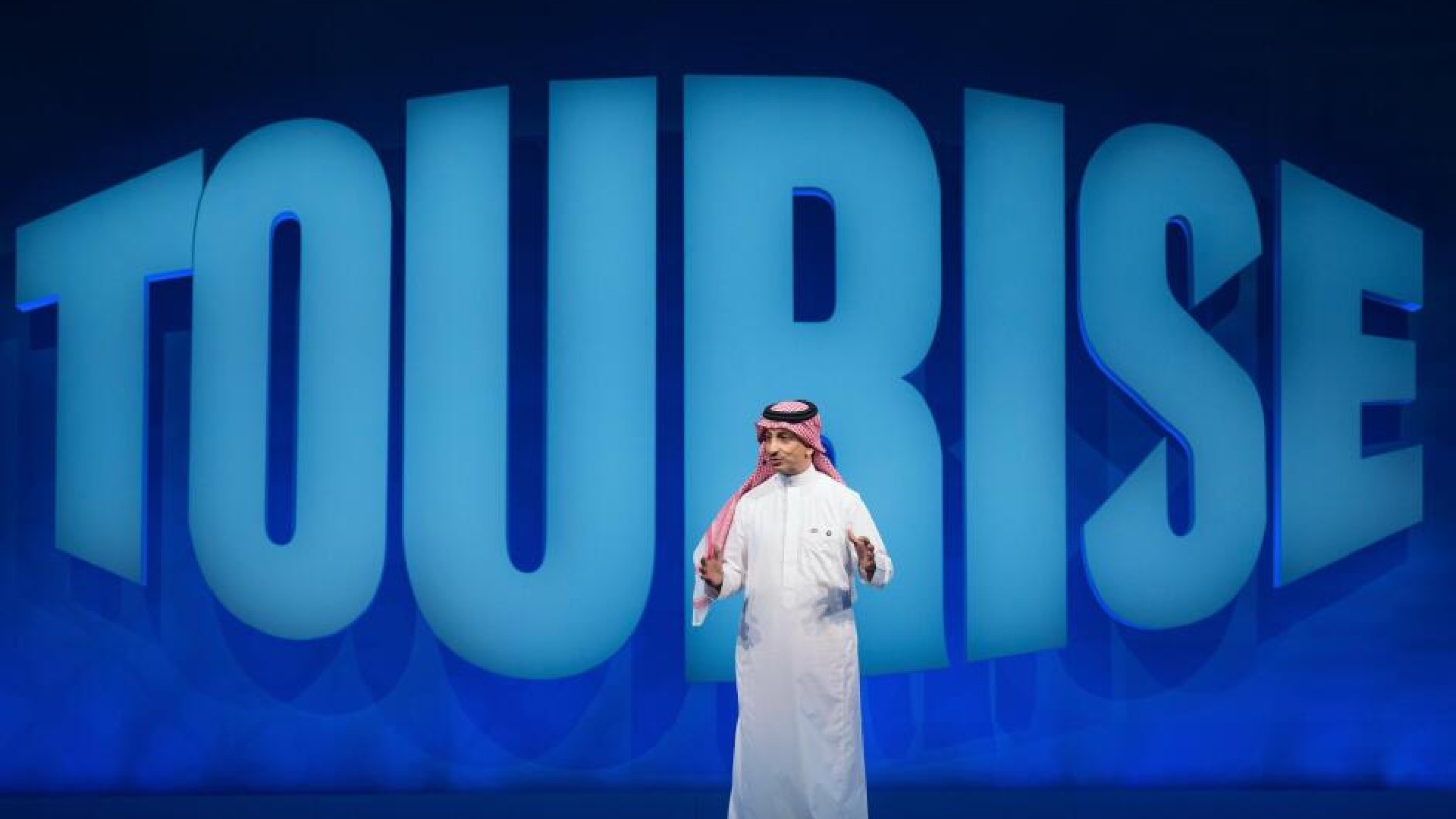 Speaker in traditional Gulf attire presenting on stage with large blue "TOURISE" text backdrop