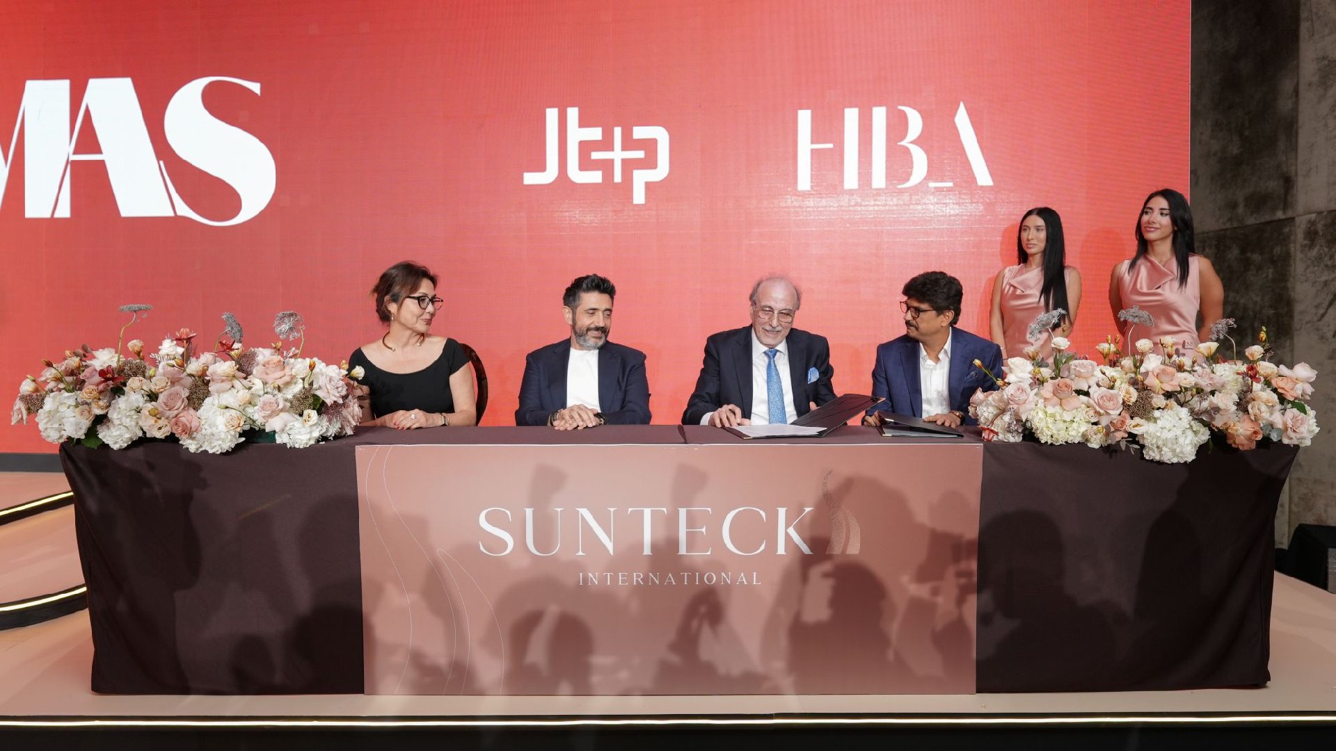 Four individuals are seated at a table covered by a cloth displaying the "Sunteck International" logo, signing documents at a ceremony, with a backdrop featuring the logos of MAS, JT+Partners, and HBA.
