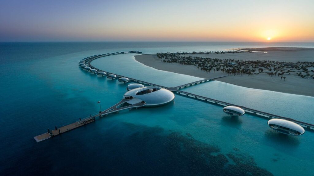 Aerial view of luxury overwater villas with curved white roofs extending into turquoise lagoon waters at sunset, with palm tree-lined beach and jetty in Saudi Arabia's Red Sea destination