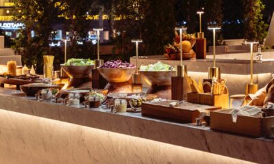 A sophisticated al-fresco dining setup on a pool terrace features a long table with a white cloth, set with wine glasses and plates, with the illuminated cityscape visible in the background.