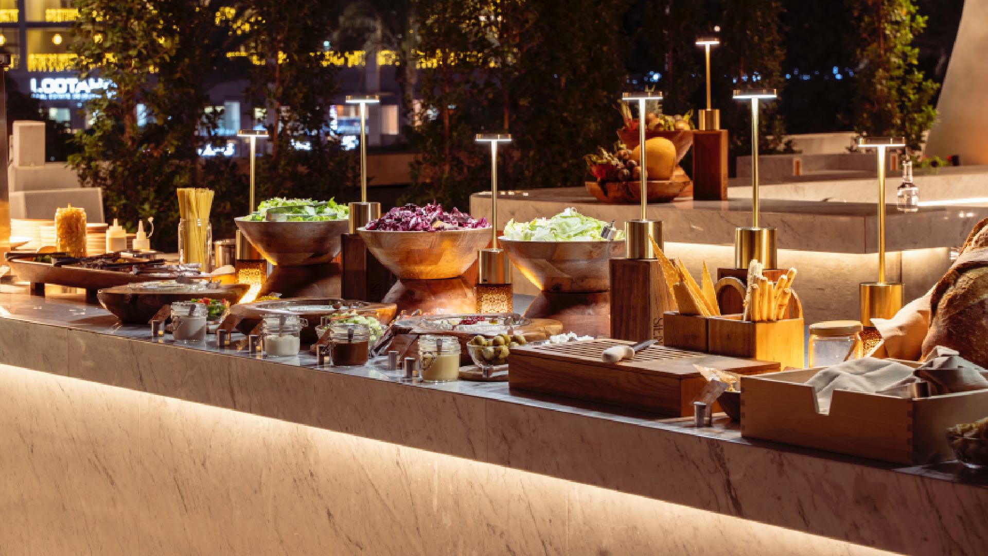 A sophisticated al-fresco dining setup on a pool terrace features a long table with a white cloth, set with wine glasses and plates, with the illuminated cityscape visible in the background.