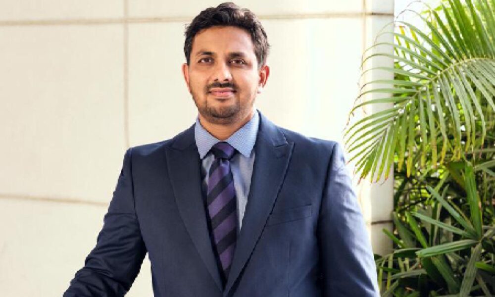 Professional portrait of a businessman in a dark suit and striped tie standing next to a green palm plant against a light background