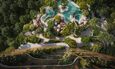 An aerial rendering shows a lush, green residential complex surrounded by forest, featuring terraced balconies covered in plants and a large, free-form swimming pool with sunbeds and white umbrellas.