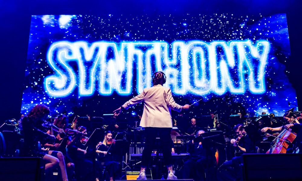 A conductor stands facing a live orchestra on stage, with the word "SYNTHONY" illuminated in bright blue neon on a large screen behind them, during a performance.