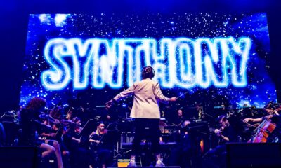 A conductor stands facing a live orchestra on stage, with the word "SYNTHONY" illuminated in bright blue neon on a large screen behind them, during a performance.