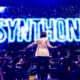 A conductor stands facing a live orchestra on stage, with the word "SYNTHONY" illuminated in bright blue neon on a large screen behind them, during a performance.