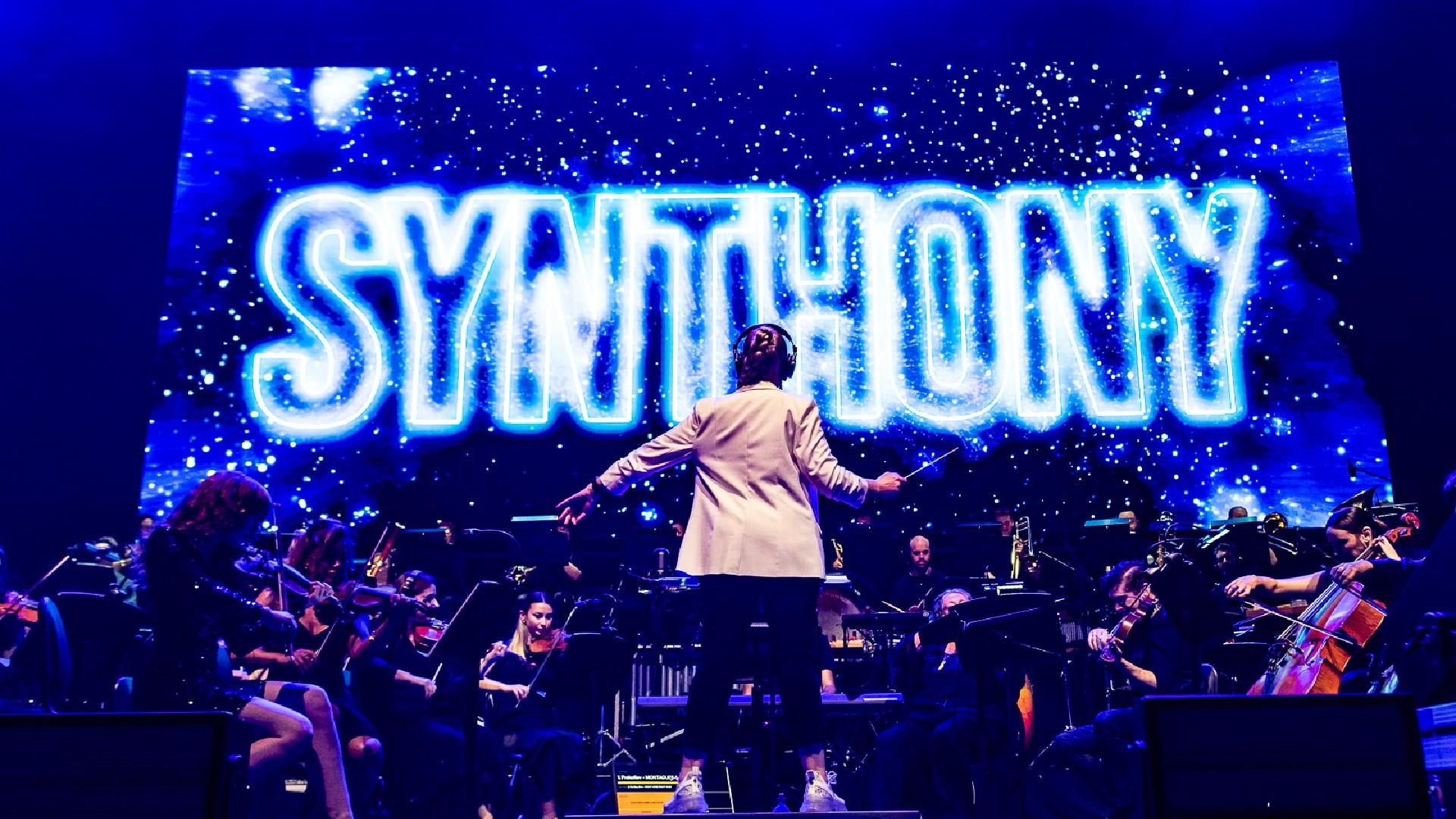 A conductor stands facing a live orchestra on stage, with the word "SYNTHONY" illuminated in bright blue neon on a large screen behind them, during a performance.