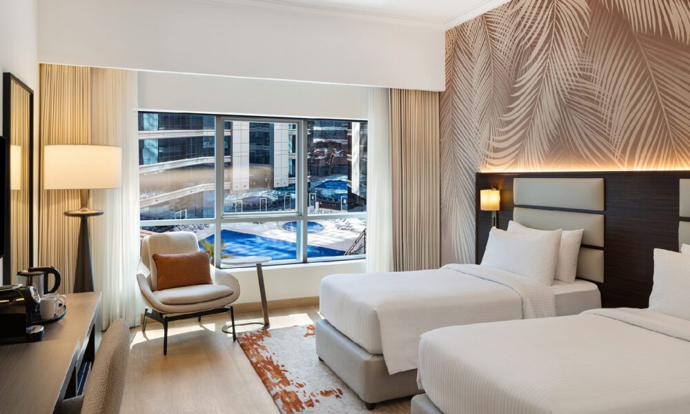 A brightly lit hotel room features two twin beds, a palm-frond patterned accent wall, and a large window overlooking a pool area and neighboring buildings.