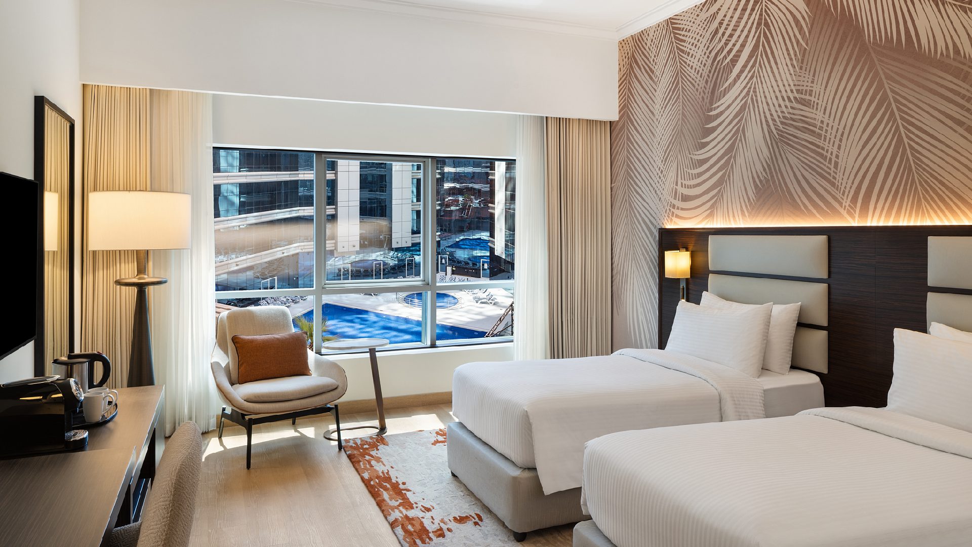 A brightly lit hotel room features two twin beds, a palm-frond patterned accent wall, and a large window overlooking a pool area and neighboring buildings.