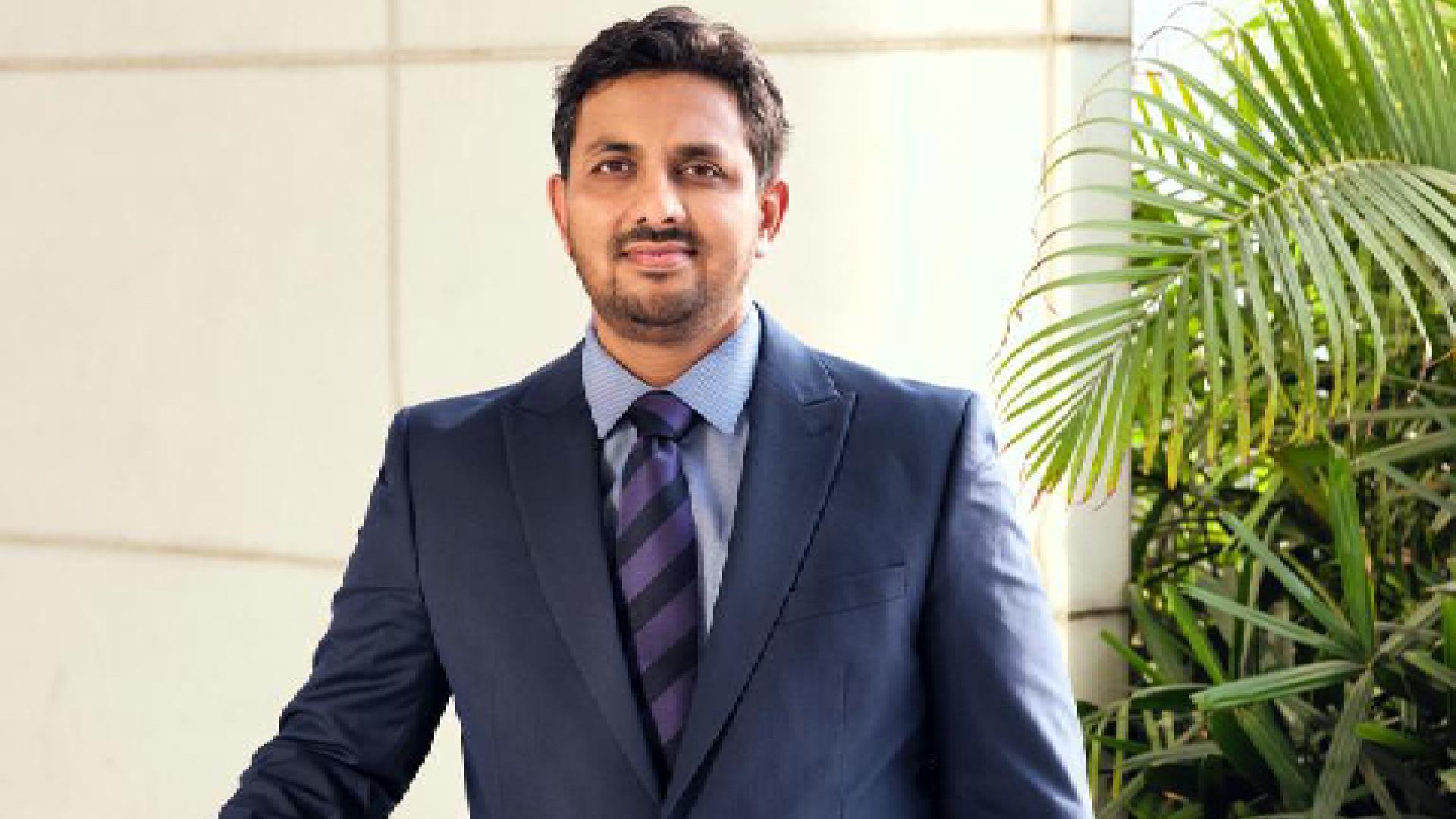 Professional portrait of a businessman in a dark suit and striped tie standing next to a green palm plant against a light background