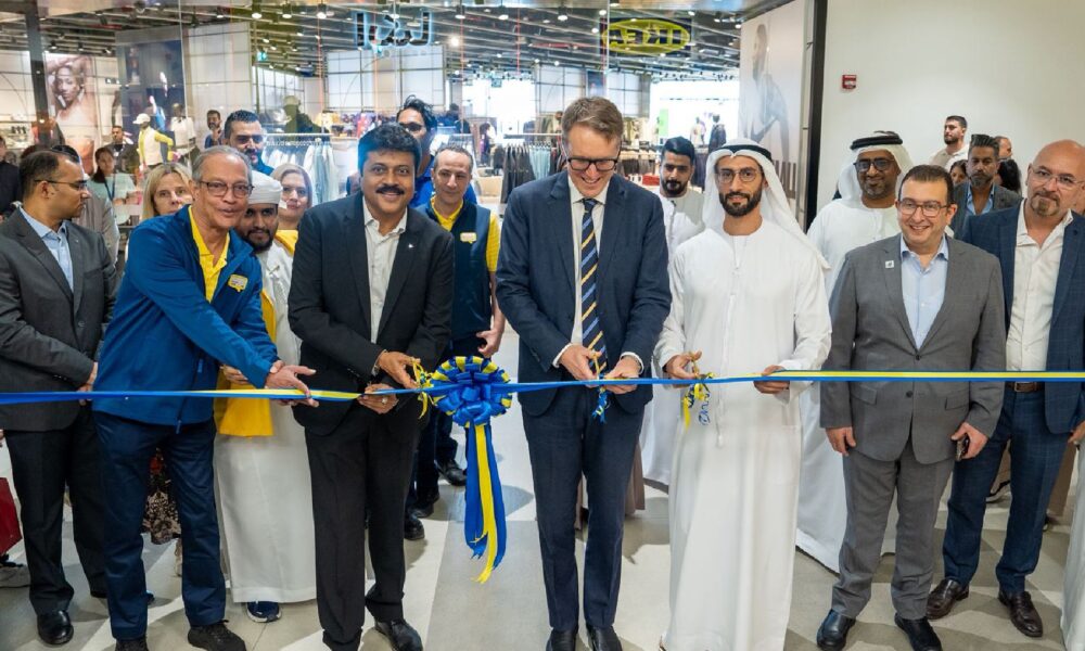 Group of business leaders and dignitaries, including men in suits and traditional white robes, cutting a blue and yellow ribbon to celebrate a store opening