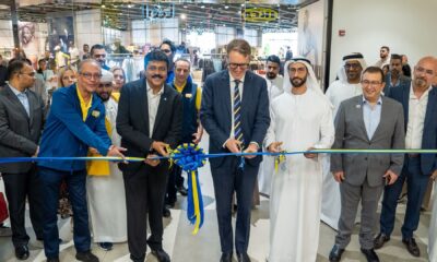 Group of business leaders and dignitaries, including men in suits and traditional white robes, cutting a blue and yellow ribbon to celebrate a store opening