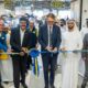 Group of business leaders and dignitaries, including men in suits and traditional white robes, cutting a blue and yellow ribbon to celebrate a store opening
