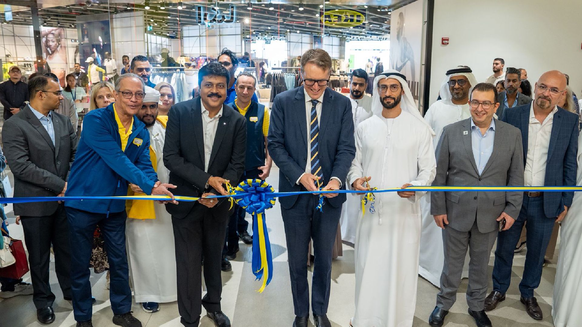 Group of business leaders and dignitaries, including men in suits and traditional white robes, cutting a blue and yellow ribbon to celebrate a store opening