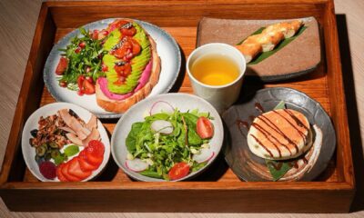 A wooden tray holding a selection of Japanese-inspired breakfast items, including avocado toast, a side salad, yogurt with granola and fruit, pancakes, and a cup of tea