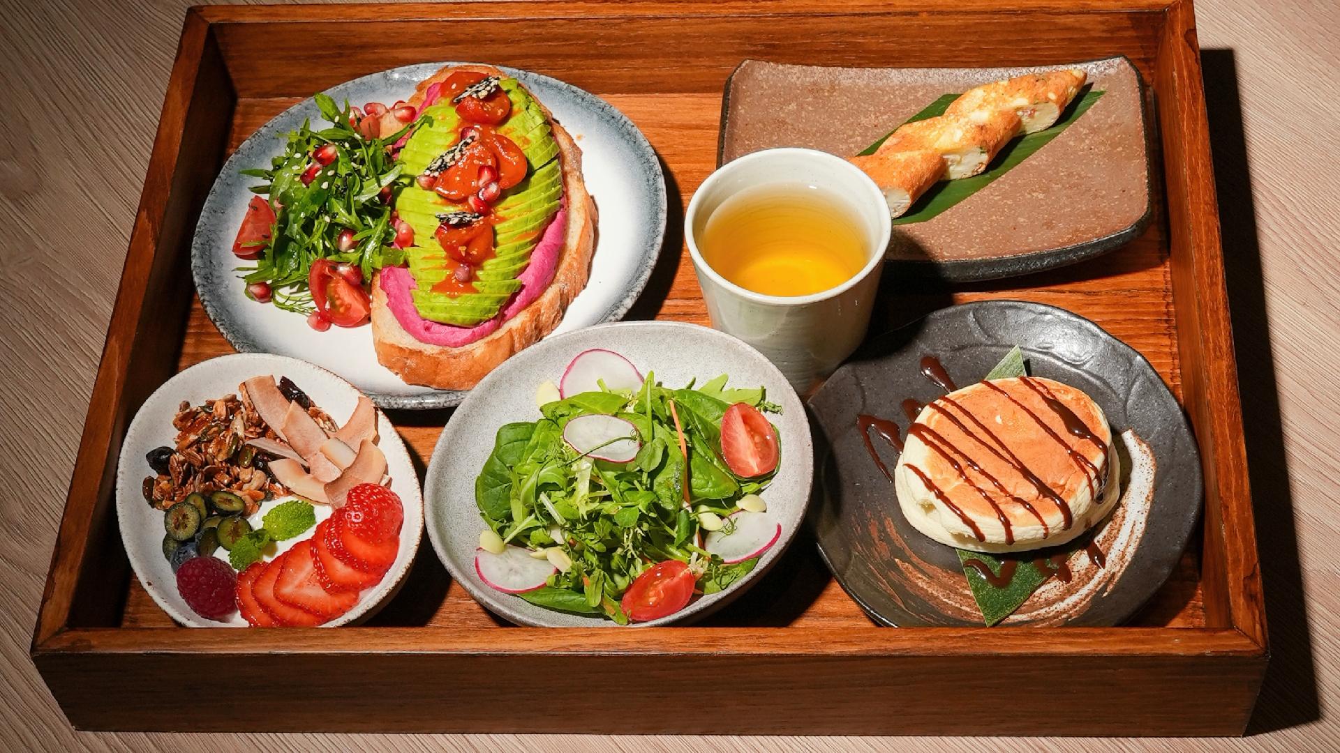 A wooden tray holding a selection of Japanese-inspired breakfast items, including avocado toast, a side salad, yogurt with granola and fruit, pancakes, and a cup of tea