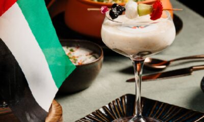A close-up shot of a creamy dessert in a stemmed glass, topped with fruit on a skewer, next to a small UAE flag