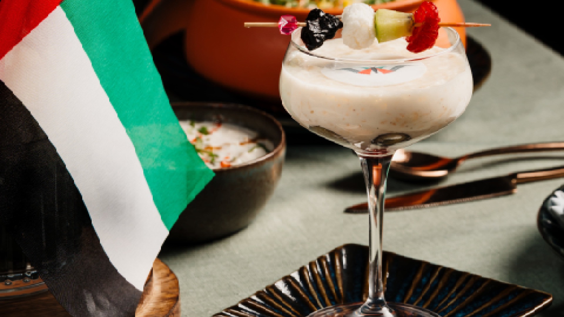 A close-up shot of a creamy dessert in a stemmed glass, topped with fruit on a skewer, next to a small UAE flag