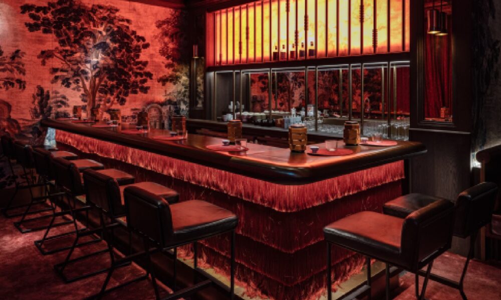 A stylized, illuminated bar area featuring a sleek, dark counter, an array of colorful liquor bottles displayed on stepped shelving, and soft accent lighting creating an intimate atmosphere.