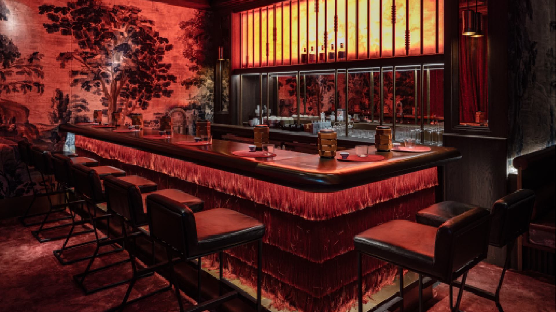 A stylized, illuminated bar area featuring a sleek, dark counter, an array of colorful liquor bottles displayed on stepped shelving, and soft accent lighting creating an intimate atmosphere.