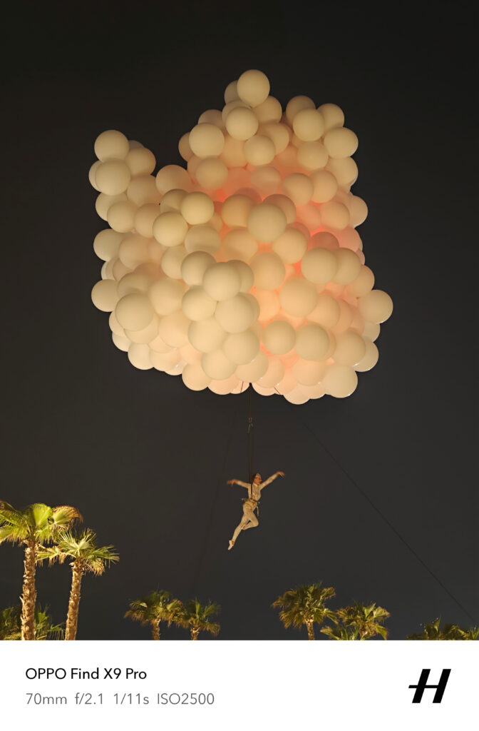 Aerial performer suspended beneath a large cluster of glowing white balloons at night, floating above palm trees during an outdoor event.