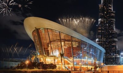 A luxurious, well-lit restaurant interior features a large white stone counter with high stools, modern minimalist decor, and a wall of geometric shelving holding decorative vases and art.