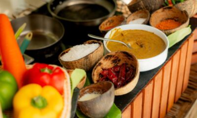 A chef is demonstrating a cooking technique over a large frying pan in an outdoor, thatched-roof cooking class area, with several Maldivian dishes laid out on a table in front of him.