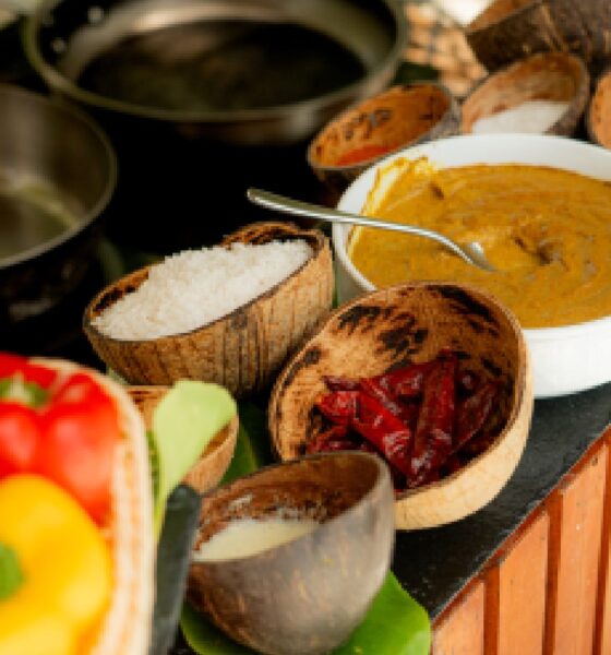 A chef is demonstrating a cooking technique over a large frying pan in an outdoor, thatched-roof cooking class area, with several Maldivian dishes laid out on a table in front of him.