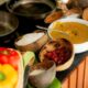 A chef is demonstrating a cooking technique over a large frying pan in an outdoor, thatched-roof cooking class area, with several Maldivian dishes laid out on a table in front of him.