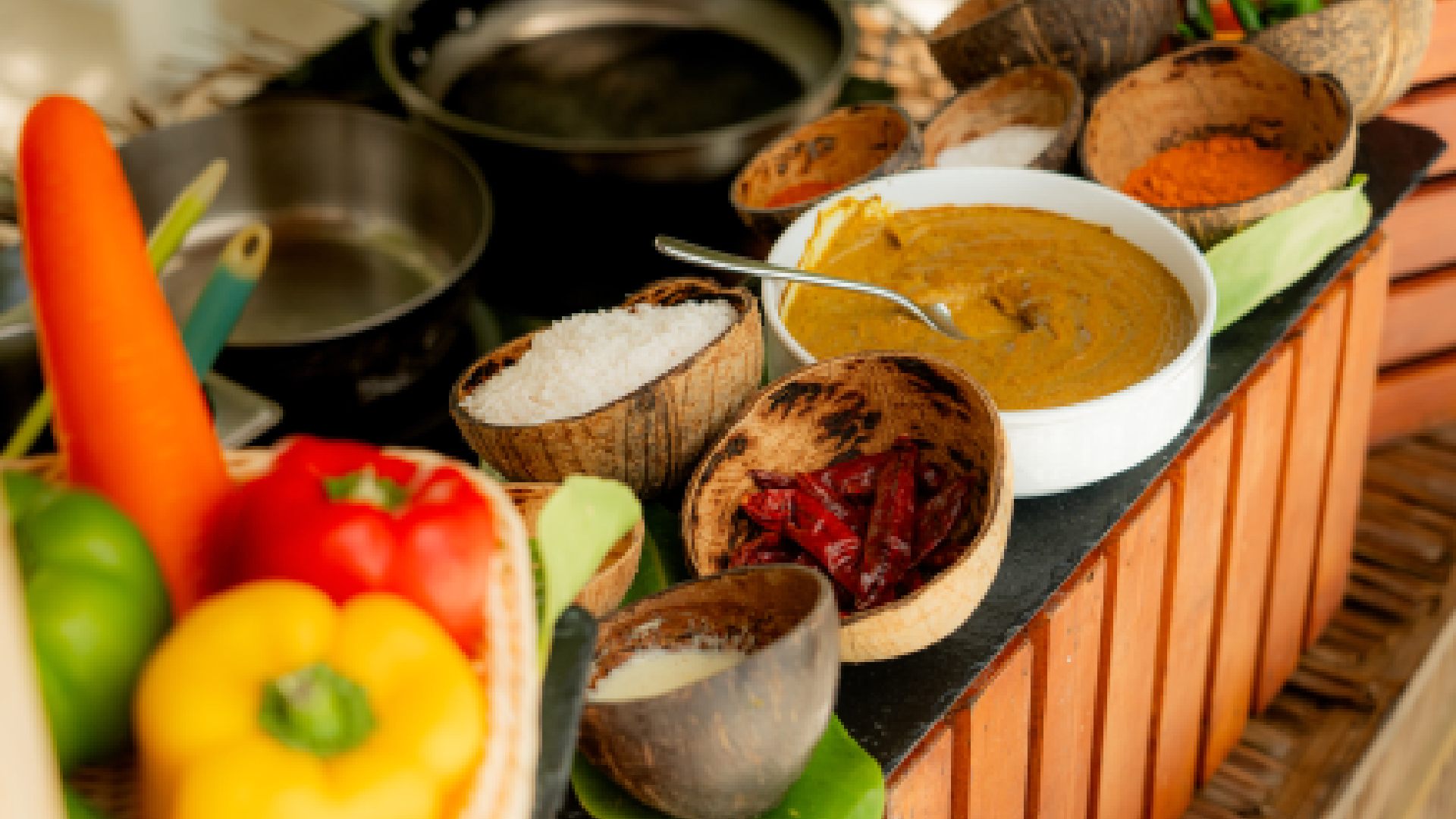 A chef is demonstrating a cooking technique over a large frying pan in an outdoor, thatched-roof cooking class area, with several Maldivian dishes laid out on a table in front of him.