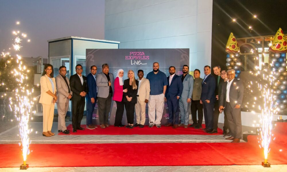 A small, minimalist desk area features a large, simple mirror, a contemporary light fixture, and a decorative vase, set against a dark-paneled wall and a white countertop.