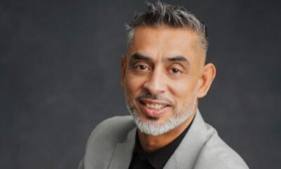 Professional headshot of a smiling businessman with grey beard and styled hair wearing a light grey blazer and black shirt against a dark grey background