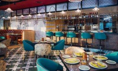 A modern Indian restaurant interior features dark wooden ceilings and seating, geometric patterned dividers, and decorative brass lanterns hanging above a row of banquette seating.