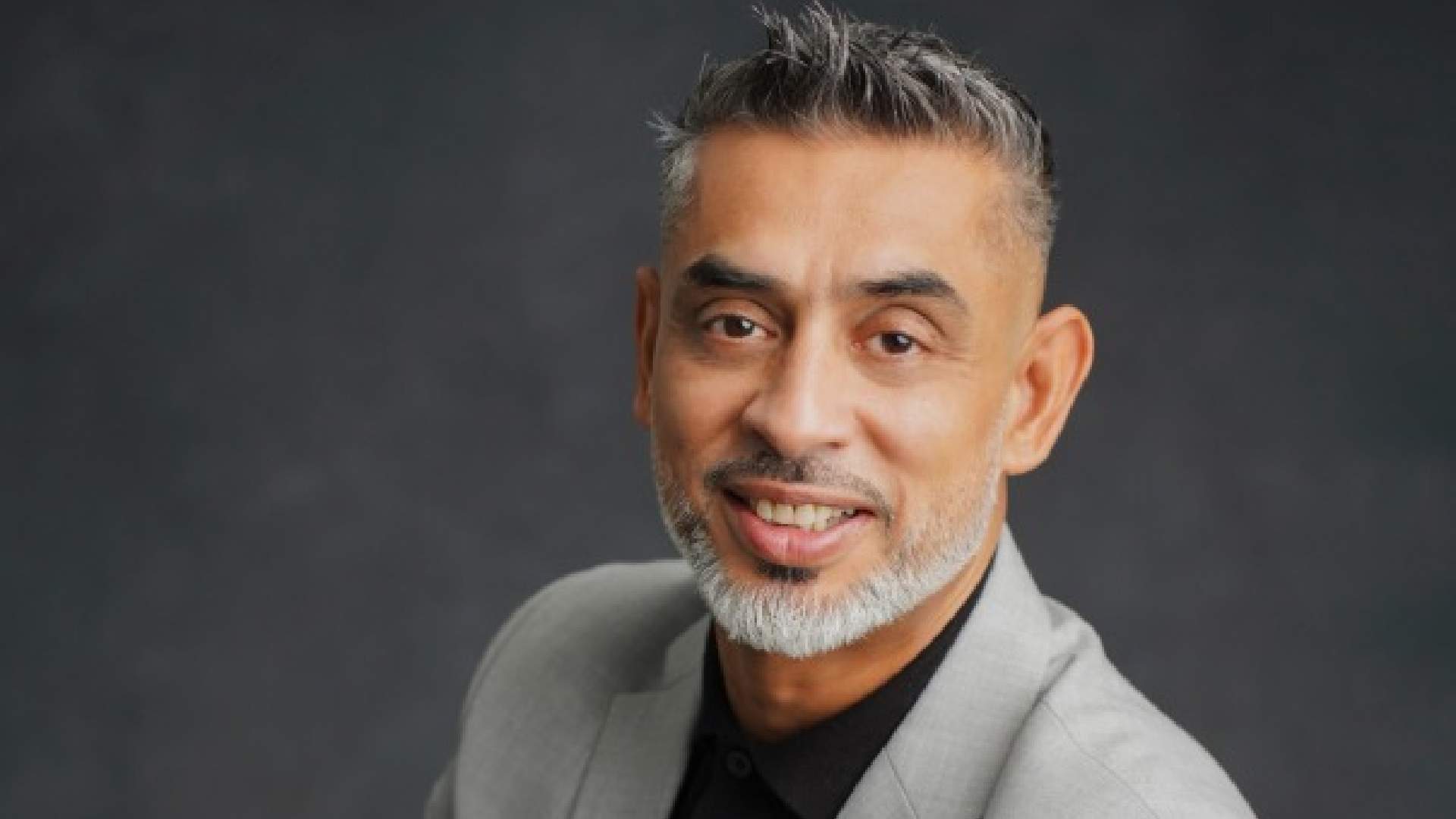 Professional headshot of a smiling businessman with grey beard and styled hair wearing a light grey blazer and black shirt against a dark grey background