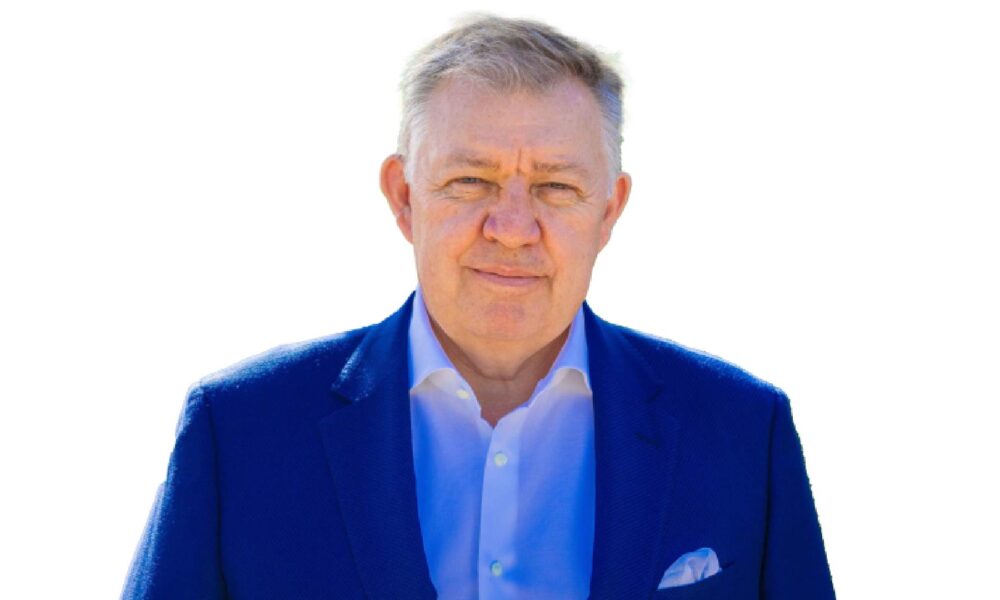 Professional headshot of a senior executive with grey hair wearing a blue blazer and light blue shirt with pocket square against a white background