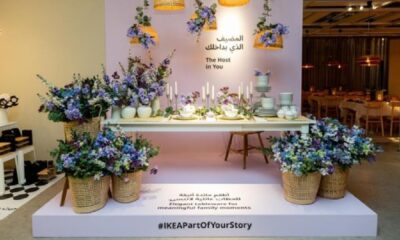 A white, modern apartment interior features a light-colored sectional sofa, a black-framed glass partition separating the living space from another room, and a decorative tree in the corner.
