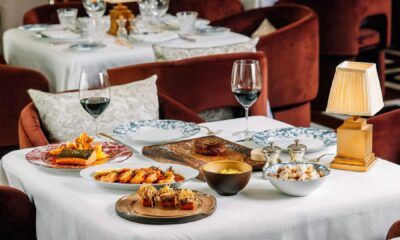 Elegant restaurant table setting with multiple gourmet dishes including steak, seafood, and appetizers, accompanied by red wine glasses and luxurious rust-colored velvet chair