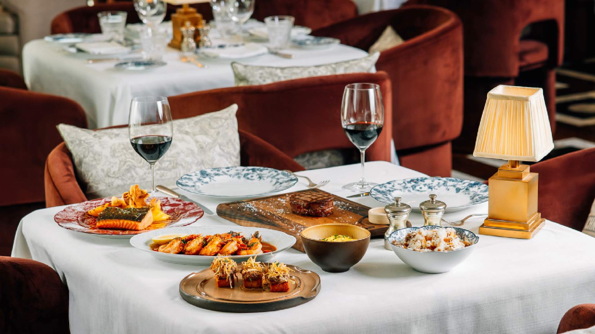 Elegant restaurant table setting with multiple gourmet dishes including steak, seafood, and appetizers, accompanied by red wine glasses and luxurious rust-colored velvet chair