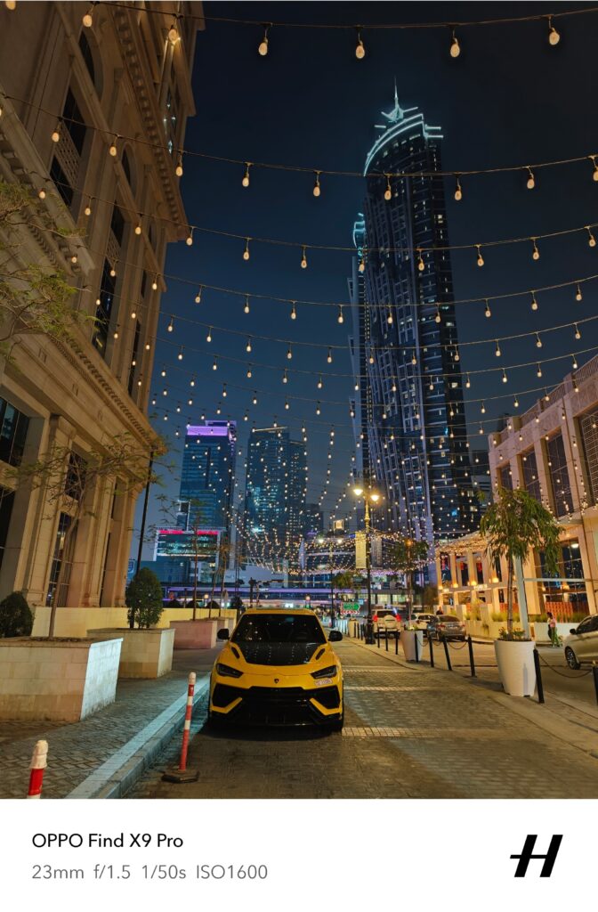 A wide angle vertical shot of a Lamborghini Urus in yellow and black color combo parked in an alleyway with high-rise Dubai building behind it and decorative lights above it.