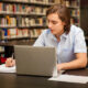 Student working on a silver laptop and taking notes with a pen in a library setting, with bookshelves visible in the background.