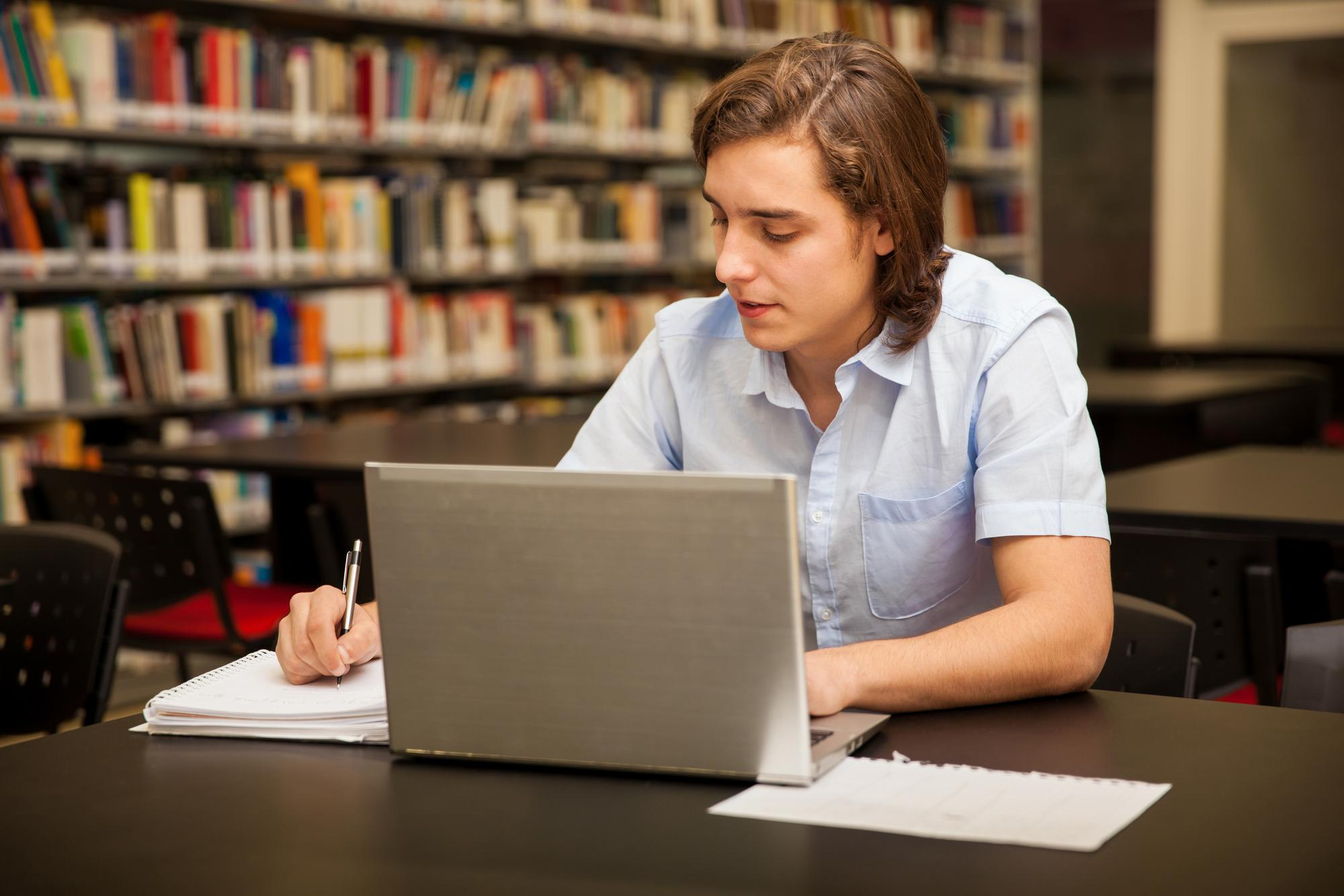 Student working on a silver laptop and taking notes with a pen in a library setting, with bookshelves visible in the background.