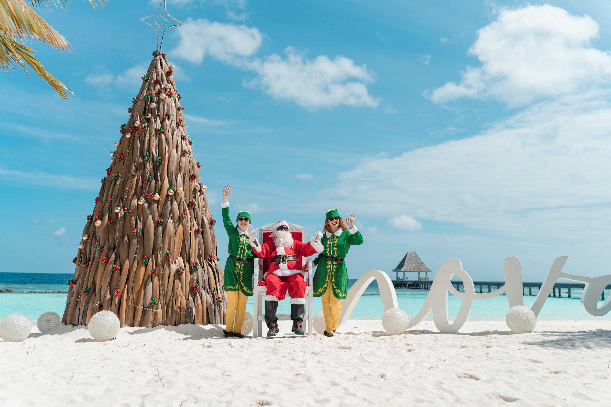 A professional lifestyle photograph at Nova Maldives during the 'Maldivian Magic' festive season, featuring a unique sustainable Christmas tree handcrafted from tiered cajon wood and driftwood.