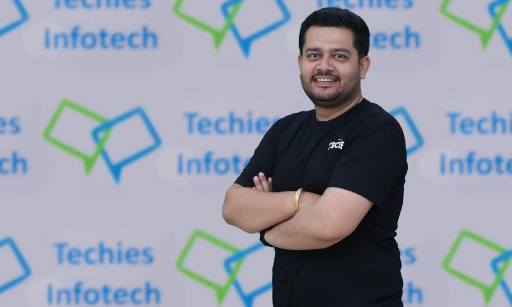 Anurag Byala, CEO, Techies Infotech, guy in black tee shirt standing infront of Techies Infotech logo