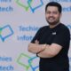 Anurag Byala, CEO, Techies Infotech, guy in black tee shirt standing infront of Techies Infotech logo