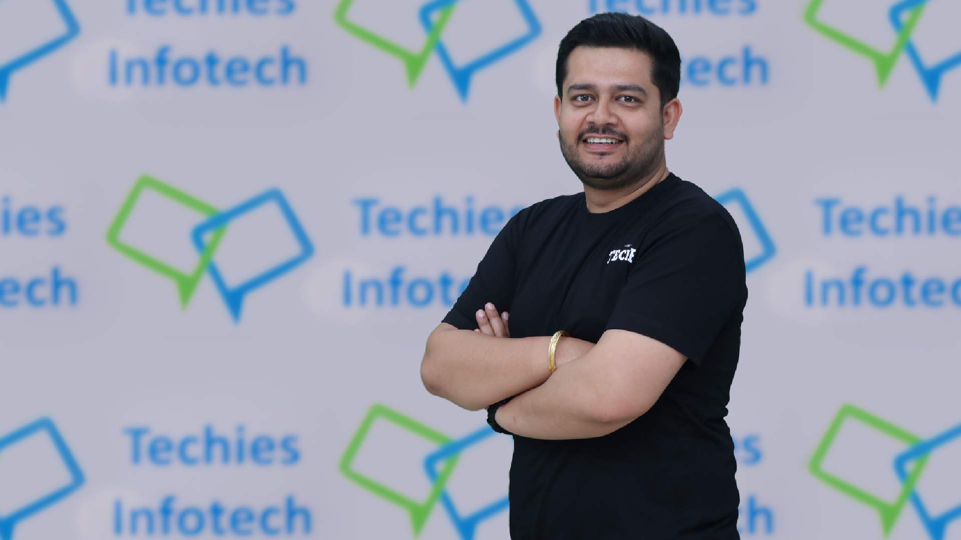 Anurag Byala, CEO, Techies Infotech, guy in black tee shirt standing infront of Techies Infotech logo