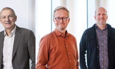 Three individuals standing indoors in front of large windows, dressed in business casual attire including a grey suit, an orange button-up shirt, and a dark jacket over a patterned shirt